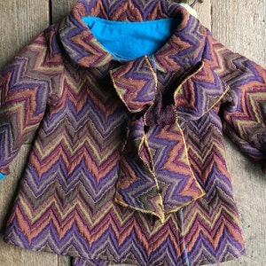 May include: A colorful baby coat with a chevron pattern in shades of purple, brown, and orange. The coat has a blue lining and cuffs, and a large bow detail. The coat is displayed on a wooden surface.