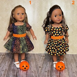 May include: Two dolls wearing Halloween-themed dresses. The first doll wears a black dress with a rainbow spiderweb pattern and an orange polka-dot waistband. The second doll wears a black dress with a candy corn print and an orange polka-dot waistband. Each doll stands next to a small orange plastic jack-o'-lantern bucket.