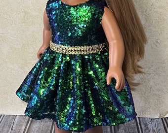 May include: A doll with long light brown hair wearing a sleeveless dress covered in shimmering green and blue sequins. The dress features a gold metallic waistband and a flared skirt. The doll is also wearing simple black flat shoes.