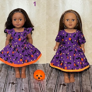 May include: Two dolls wearing purple dresses with a Halloween print featuring black bats, orange pumpkins, and yellow stars. Doll 1 has flutter sleeves and an orange hem. Doll 2 has short sleeves, an orange trim, and a black lace hem detail.
