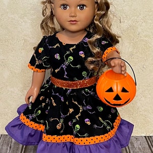 May include: A doll with wavy brown hair wearing a black dress featuring a colorful skeleton print, an orange glitter waistband, and a purple ruffled hem with orange polka dot trim, holding a small orange plastic jack-o'-lantern candy bucket.