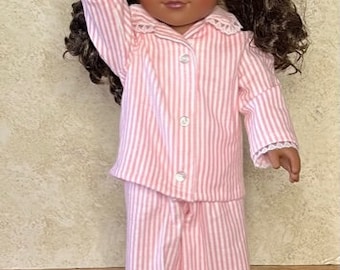 May include: A doll wearing a pink and white striped pajama set. The doll has brown hair in pigtails and is raising one arm. The pajama top has a collar and buttons down the front. The pajama pants are wide-legged.