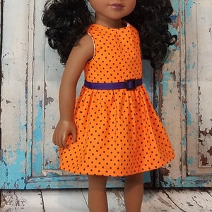 May include: A doll with dark skin and black hair styled in two pigtails. The doll is wearing an orange sleeveless dress featuring a pattern of small black polka dots and a purple waistband with a small black buckle at the center of the waist.