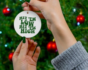 Op de afbeelding: Een witte keramische kerstbal met de tekst "Hit 'em Low Hit 'em High" in groene letters. De bal is rond en heeft een gouden touwtje om op te hangen. De achtergrond is een kerstboom met rode ballen.