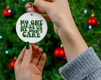 Op de afbeelding: Een witte keramische kerstbal met de tekst "NO ONE LIKES US WE DON'T CARE" in groene letters. De ronde ornament heeft een gouden touwtje om op te hangen. De achtergrond is een kerstboom met rode ballen.