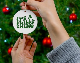 Op de afbeelding: Een witte, ronde kerstbal met de groene tekst "IT'S A PHILLY THING". De ornament wordt vastgehouden voor een kerstboom versierd met rode ballen.