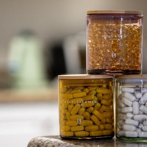 May include: Three glass jars with wooden lids stacked on top of each other, filled with supplements. The top jar contains collagen capsules, the middle jar has multivitamin tablets, and the bottom jar is filled with calcium capsules.