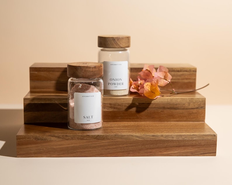 May include: Two glass spice jars with wooden lids labeled "Salt" and "Onion Powder" sit on a tiered wooden display. A sprig of dried orange and pink flowers adds a decorative touch. The jars are filled with pink salt and white powder.
