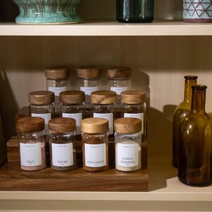 May include: A wooden spice rack displays glass jars with wooden lids, labeled with spice names like "Salt," "Thyme," and "Cinnamon." Amber glass bottles are arranged on the shelf. The jars are filled with spices and are ready to be used in the kitchen.