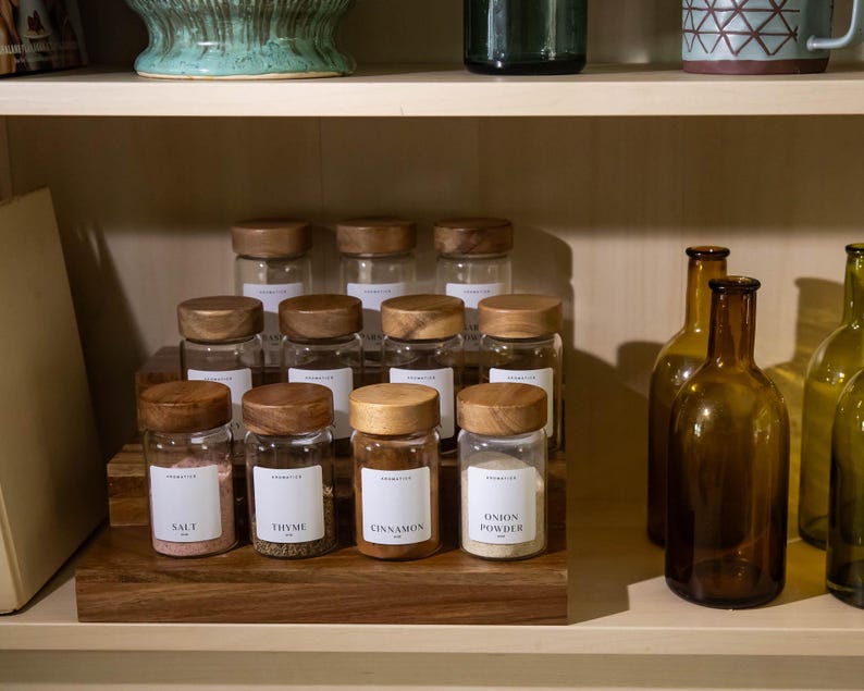 May include: A wooden spice rack displays glass jars with wooden lids, labeled with spice names like "Salt," "Thyme," and "Cinnamon." Amber glass bottles are arranged on the shelf. The jars are filled with spices and are ready to be used in the kitchen.