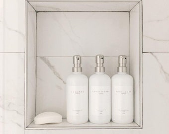 May include: Three white bottles with silver pumps in a white tiled shower recess. The bottles are labelled "Shampoo", "Conditioner", and "Body Wash". A white bar of soap is on the shelf below the bottles.