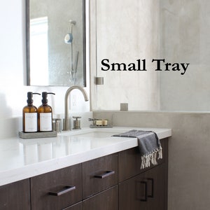 Peut inclure: Une vasque de salle de bain avec un plan de travail blanc et des armoires brun fonc&eacute;. Le plan de travail comporte un petit plateau gris avec deux flacons en verre ambr&eacute; avec pompes et une barre de savon. Le robinet est chrom&eacute; et une serviette est accroch&eacute;e &agrave; la porte de l'armoire. Le texte "Small Tray" est visible sur le mur.