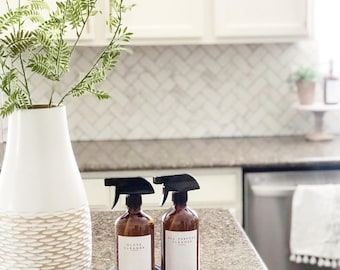 May include: Two amber glass spray bottles with black spray tops and white labels. The labels read "Glass Cleaner" and "All Purpose Cleaner". The bottles are sitting on a blue and white striped towel on a brown countertop.