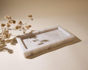 May include: A rectangular white marble tray with subtle grey veining and a smooth, polished finish. The tray features a recessed center and raised edges, accompanied by a small cluster of dried, light brown wildflowers.