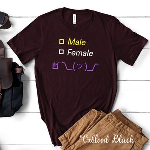 May include: Oxblood black t-shirt with yellow and white text that reads "Male" and "Female" with a checkmark and a purple emoticon. The shirt is next to a brown leather bag and tan pants.