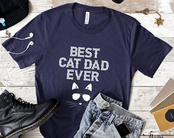 May include: A navy blue t-shirt with the text "Best Cat Dad Ever" in white. A white cat face with sunglasses is below the text. The shirt is laid flat on a woodgrain surface with a black beanie, white earbuds, black boots, blue jeans, sunglasses, and a key chain.