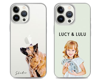 Puede incluir: Dos fundas transparentes para teléfono con diseños diferentes. La funda de la izquierda presenta una ilustración de acuarela de un perro marrón y tostado con el nombre "Shadow" escrito debajo. La funda de la derecha presenta una ilustración de acuarela de una niña con cabello rubio sosteniendo un conejo gris. El texto "LUCY & LULU" está impreso encima de la imagen.