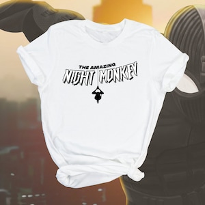 Op de afbeelding: Wit T-shirt met een zwarte grafische print van een persoon die ondersteboven hangt met de tekst "The Amazing Night Monkey" in een vet, zwart lettertype.
