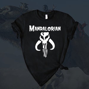 Puede incluir: Camiseta negra con un s&iacute;mbolo mandaloriano blanco y el texto "Mandalorian" impreso en la parte delantera.
