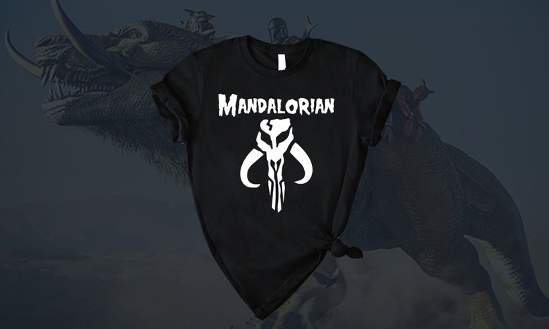 Puede incluir: Camiseta negra con un s&iacute;mbolo mandaloriano blanco y el texto "Mandalorian" impreso en la parte delantera.