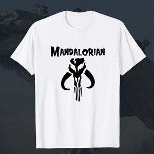 Puede incluir: Camiseta blanca con un s&iacute;mbolo mandaloriano negro y el texto "MANDALORIAN" impreso en la parte delantera.