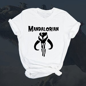 Puede incluir: Camiseta blanca con un s&iacute;mbolo mandaloriano negro y el texto "MANDALORIAN" impreso en la parte delantera.
