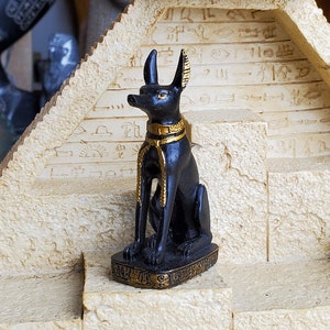 Op de afbeelding: Een klein zwart Anubis-beeldje met gouden accenten staat op een beige piramidevormige displaystandaard. De standaard heeft een getextureerd oppervlak en is versierd met hiërogliefen.