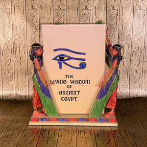 May include: A decorative figurine featuring two Egyptian women flanking a white frame with a blue Eye of Horus symbol and the text "The Living Wisdom of Ancient Egypt".