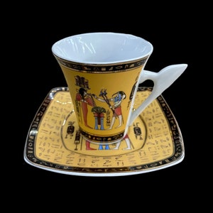 Könnte beinhalten: Eine weiße Teetasse und Untertasse aus Keramik mit gelbem Hintergrund und schwarzen und goldenen Akzenten. Tasse und Untertasse zeigen ein ägyptisch inspiriertes Design mit Hieroglyphen und Figuren.