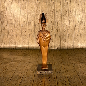 May include: A gold-colored statue of an Egyptian pharaoh standing on a base with hieroglyphics. The statue is in front of a wall with hieroglyphics.