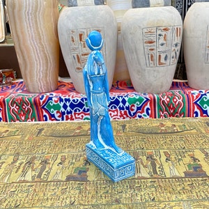 Puede incluir: Una figurita de cerámica azul de una diosa egipcia de pie sobre una base con jeroglíficos. La figurita está delante de una mesa con una imagen impresa de jeroglíficos.