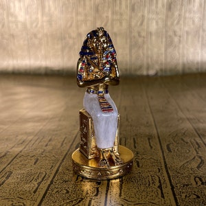 May include: A gold-colored bust of an Egyptian pharaoh with a blue and red headdress. The bust sits atop a white and gold pedestal, with a gold base. The background is a gold surface with hieroglyphs.