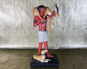 May include: A figurine of an Egyptian man carrying a gazelle on his shoulders. He is holding a smaller gazelle in his left hand and a dog is at his feet. The figurine is on a black base.