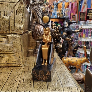 May include: Gold and black figurine of an Egyptian goddess sitting on a throne. The goddess is wearing a crown and holding a small figure in her lap. The figurine is on a black base with a gold design.