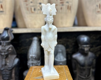 May include: White figurine of an Egyptian pharaoh wearing a crown and a kilt. The figurine is standing on a gold-colored base. The figurine is in front of a shelf with several white vases with hieroglyphics.