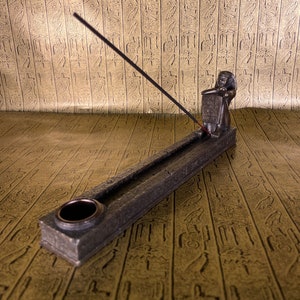 May include: A black incense stick burning in a gray stone incense holder shaped like an Egyptian pharaoh holding a tablet. The holder is on a gold patterned surface.
