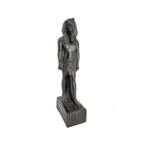 May include: Black statue of an Egyptian pharaoh standing on a base with hieroglyphics. The statue is made of a dark, textured material and has a detailed design.