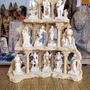 May include: A set of 12 white porcelain figurines of Greek gods and goddesses, arranged in a tiered display on a stone-like base. The figurines are painted in gold and white, and each has a name inscription below.