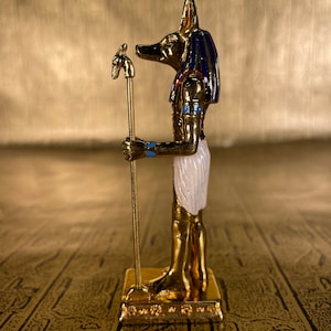 May include: A gold figurine of the Egyptian god Anubis, depicted as a jackal with a human body, holding a staff with a jackal head. The figurine is standing on a gold base and has blue and white details.