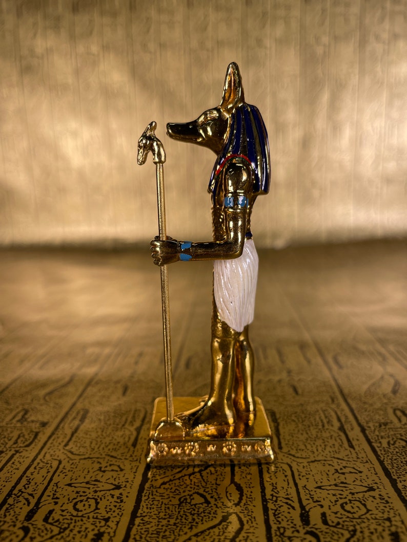 May include: A gold figurine of the Egyptian god Anubis, depicted as a jackal with a human body, holding a staff with a jackal head. The figurine is standing on a gold base and has blue and white details.
