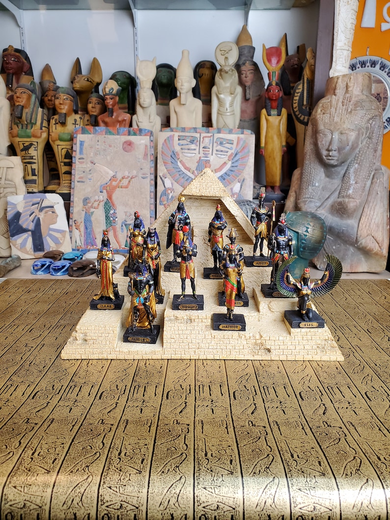May include: A set of 10 Egyptian god figurines, each standing on a small platform with their name printed below. The figurines are arranged in a pyramid shape on a base with hieroglyphics.