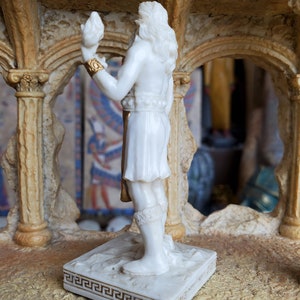 May include: White marble statue of a figure holding a bird in their hand. The statue is standing on a square base with a gold Greek key design.