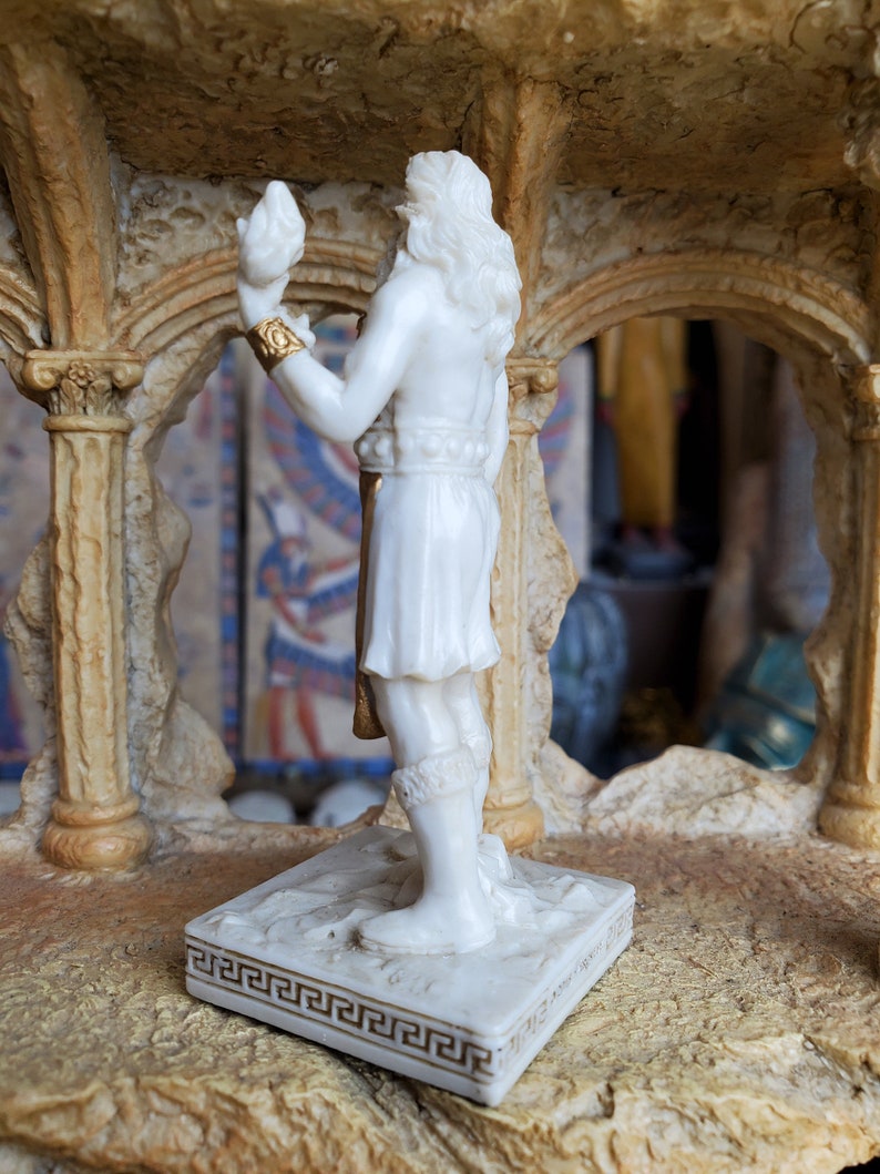 May include: White marble statue of a figure holding a bird in their hand. The statue is standing on a square base with a gold Greek key design.