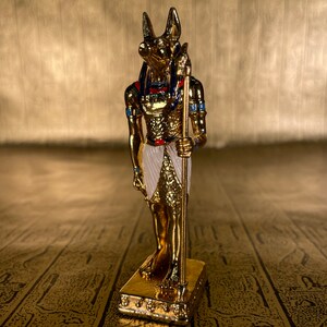 May include: A gold figurine of the Egyptian god Anubis, standing on a gold base. Anubis is depicted as a jackal-headed man holding a staff.