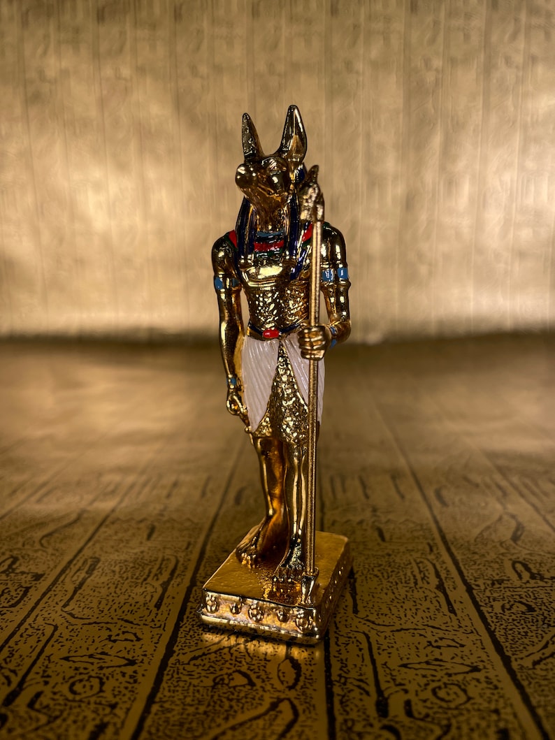 May include: A gold figurine of the Egyptian god Anubis, standing on a gold base. Anubis is depicted as a jackal-headed man holding a staff.