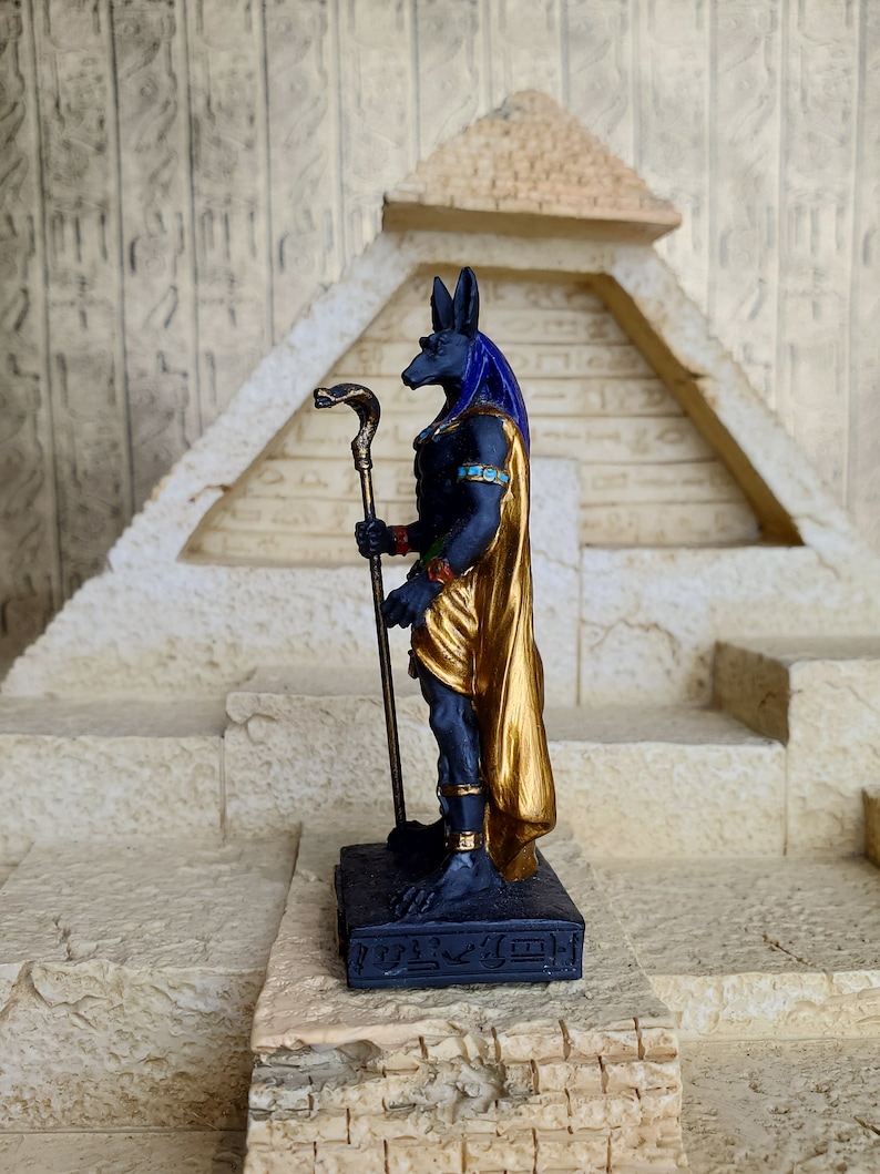 May include: Black and gold figurine of the Egyptian god Anubis standing on a base with hieroglyphics. Anubis is depicted as a jackal-headed man holding a staff with a snake head.