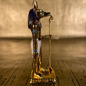 May include: A gold-colored figurine of the Egyptian god Anubis, with a jackal head, standing on a base. The figurine is painted with blue, white, and gold accents.