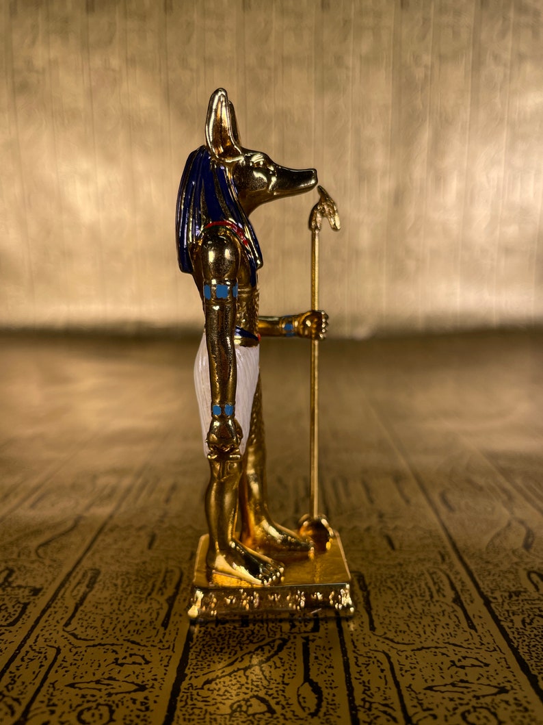 May include: A gold-colored figurine of the Egyptian god Anubis, with a jackal head, standing on a base. The figurine is painted with blue, white, and gold accents.
