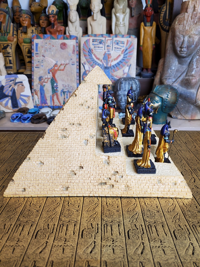 May include: A miniature pyramid with a beige stone finish and a row of gold and blue Egyptian figurines standing on a platform. The pyramid is on a surface with a pattern of hieroglyphics.
