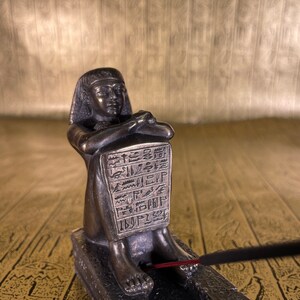 May include: Bronze incense burner shaped like an Egyptian pharaoh sitting with crossed legs. The burner has a rectangular incense holder with hieroglyphics carved into it. A stick of incense is burning in the holder.